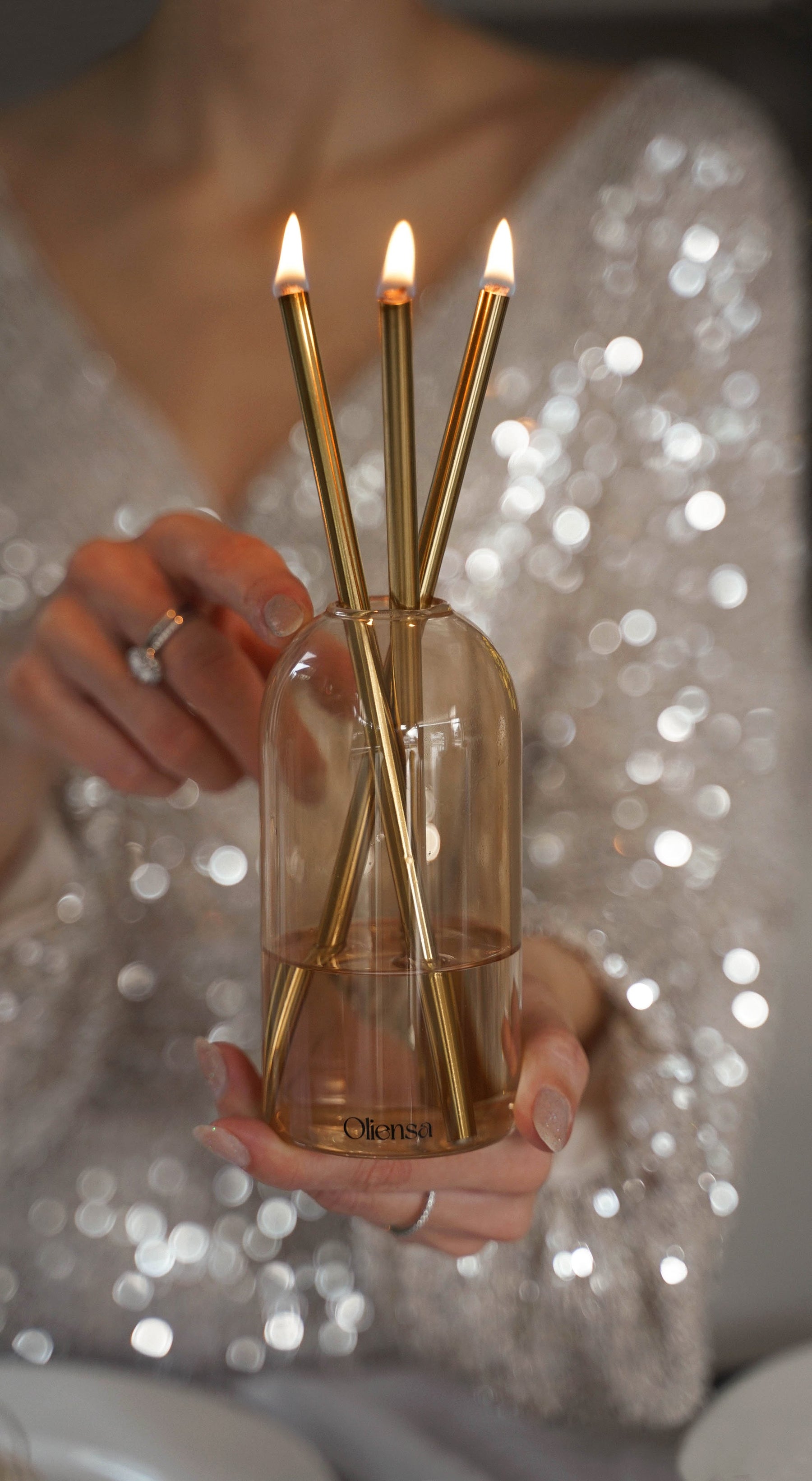 Ekskluzywna świeca oliensa eternal gold sand, trzymana przez bardzo elegancką kobietę w cekinowej sukni. Zbliżenie na detale świecy raz zadbane dłonie kobiety.