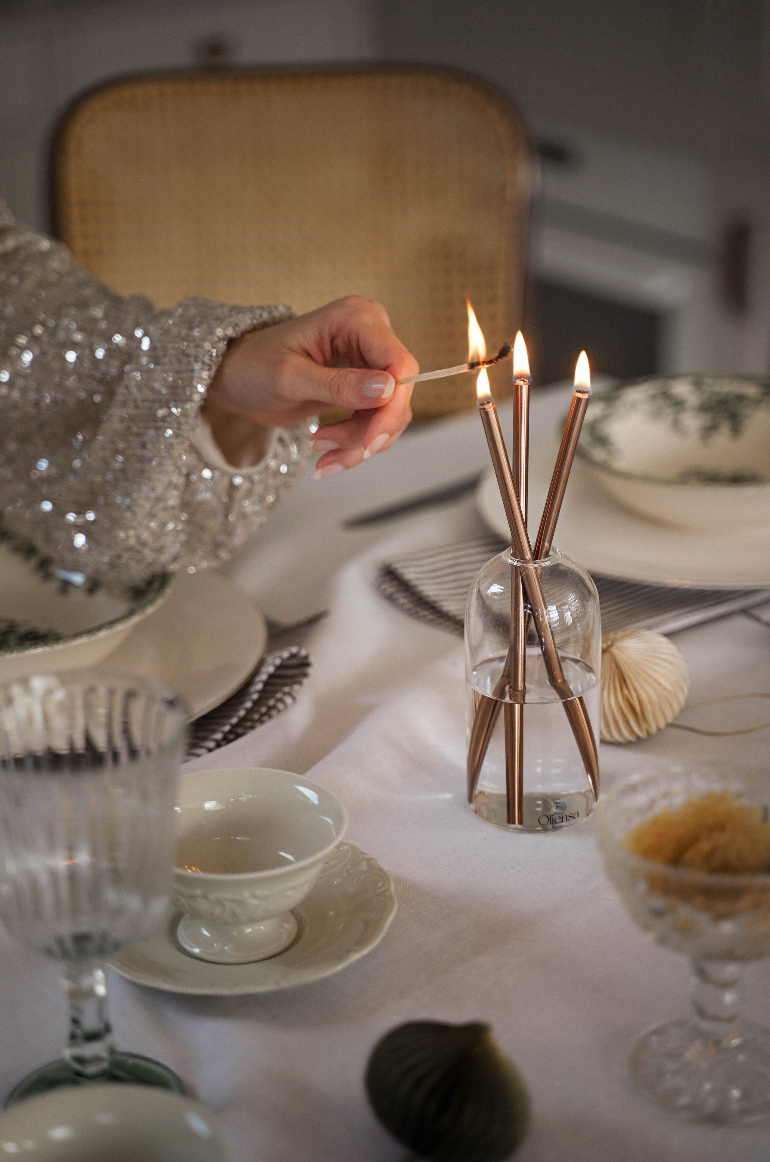 Stół nakryty porcelaną oraz delikatna kobieca dłoń, podpalające metalowe świece Oliensa Eternal Copper Crystal. 